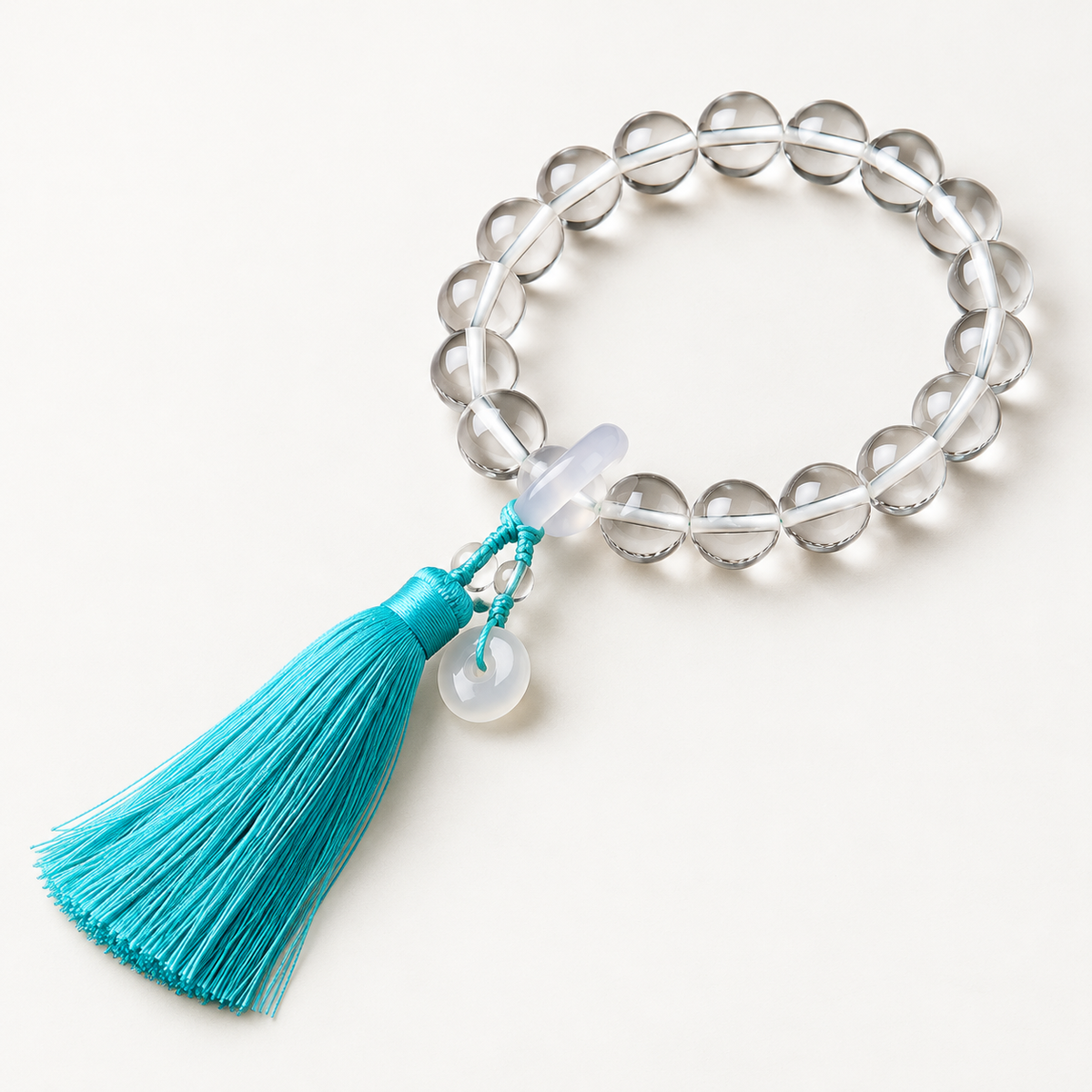 18 transparent round main beads with pale ring details and a teal cord tassel on an off-white background.